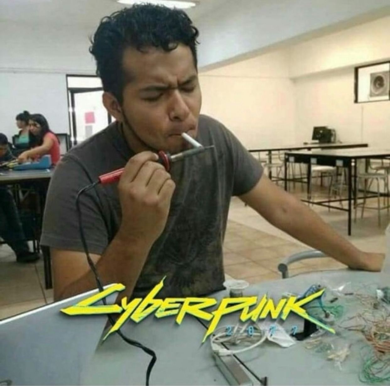 This meme is a picture of a person with a confused expression, sitting at a desk with a computer and various electronics scattered around them. The image is captioned "Cyberpunk 2077" and features the game's logo, which is a stylized, futuristic font. The meme is likely a humorous commentary on the game's notorious bugs and technical issues, with the image implying that the person is struggling to fix a problem with their computer.
